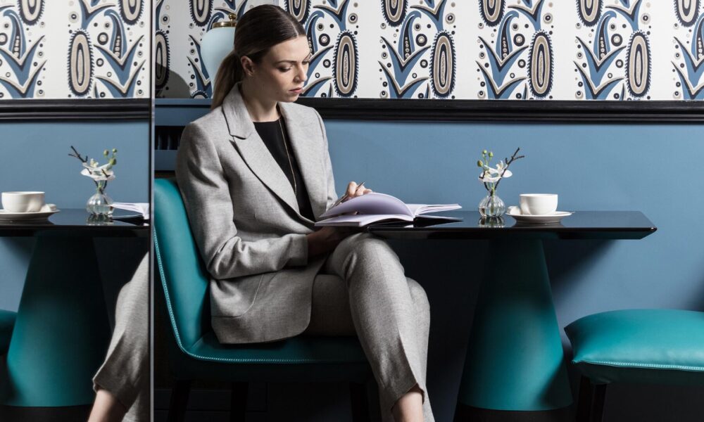 Mulher elegante de fato cinzento sentada no lounge do Hotel Malte, com paredes azuis e papel de parede gráfico ao fundo