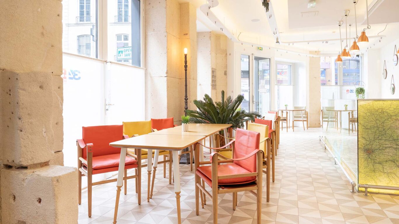 Vue de la Salle Marianne de l’hôtel 34B, un espace lumineux aux grandes fenêtres, agrémenté de tables en bois et de chaises rouges et jaunes pour une atmosphère conviviale et colorée.