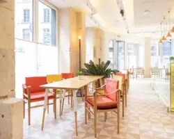 Vue de la Salle Marianne de l’hôtel 34B, un espace lumineux aux grandes fenêtres, agrémenté de tables en bois et de chaises rouges et jaunes pour une atmosphère conviviale et colorée.