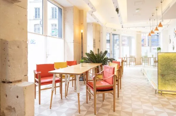 Vue de la Salle Marianne de l’hôtel 34B, un espace lumineux aux grandes fenêtres, agrémenté de tables en bois et de chaises rouges et jaunes pour une atmosphère conviviale et colorée.