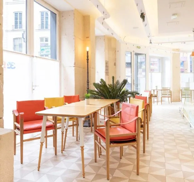 Vue de la Salle Marianne de l’hôtel 34B, un espace lumineux aux grandes fenêtres, agrémenté de tables en bois et de chaises rouges et jaunes pour une atmosphère conviviale et colorée.