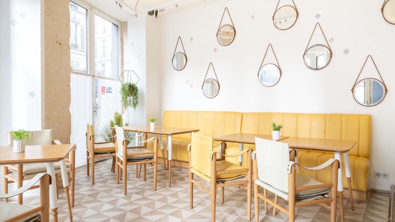 Vue de la Salle Marianne de l’hôtel 34B, un espace lumineux aux grandes fenêtres, agrémenté de tables en bois et de chaises rouges et jaunes pour une atmosphère conviviale et colorée.