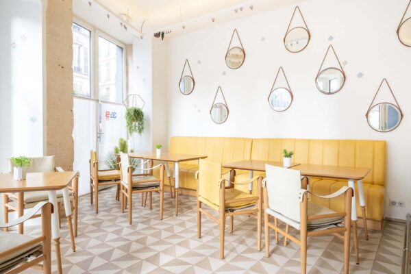 Vue de la Salle Marianne de l’hôtel 34B, un espace lumineux aux grandes fenêtres, agrémenté de tables en bois et de chaises rouges et jaunes pour une atmosphère conviviale et colorée.