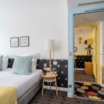 Chambre de l’hôtel Augustin à Paris avec lit double, coussins décoratifs et vue sur une seconde pièce par une porte ouverte