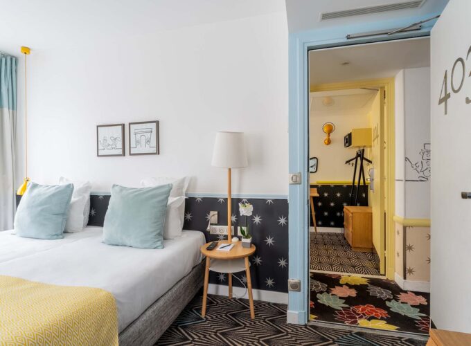 Chambre de l’hôtel Augustin à Paris avec lit double, coussins décoratifs et vue sur une seconde pièce par une porte ouverte