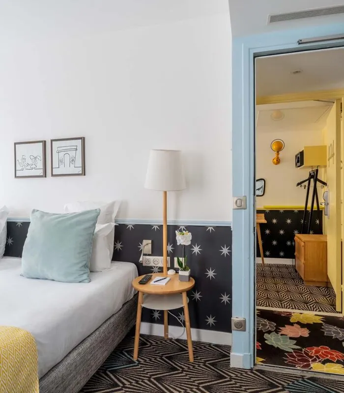 Chambre de l’hôtel Augustin à Paris avec lit double, coussins décoratifs et vue sur une seconde pièce par une porte ouverte
