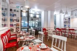 Salle de petit-déjeuner de l’hôtel 3 étoiles Monterosa du groupe Astotel à Paris, avec des chaises rouges et blanches, des tables en bois et une décoration murale sur le thème des livres