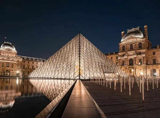 La pyramide de verre du musée du Louvre à Paris, éclairée de nuit, avec le palais historique en arrière-plan.
