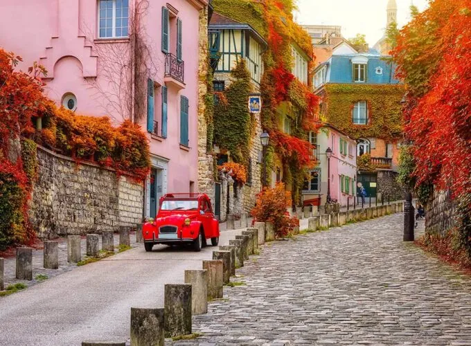 Ruelle pavée de Montmartre à Paris en automne, avec une voiture ancienne rouge (2CV), des maisons anciennes et des façades couvertes de vigne vierge.