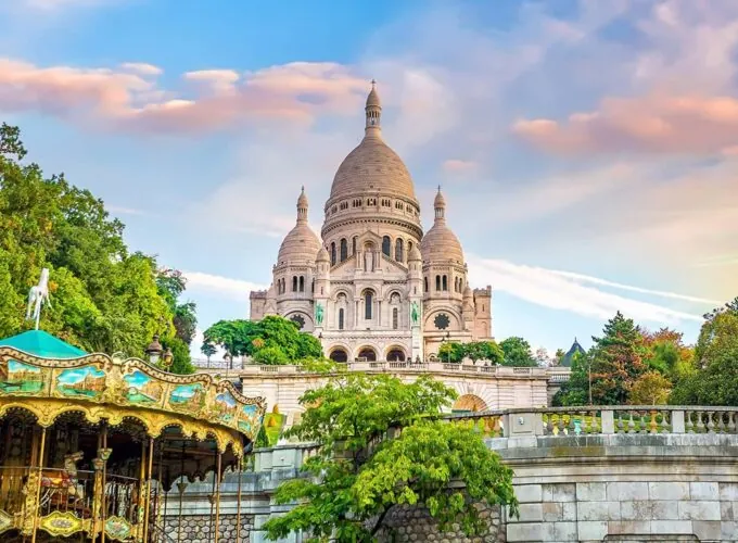 Vue sur la basilique du Sacré-Cœur de Montmartre à Paris au coucher du soleil, avec un carrousel en premier plan et des arbres verdoyants.
