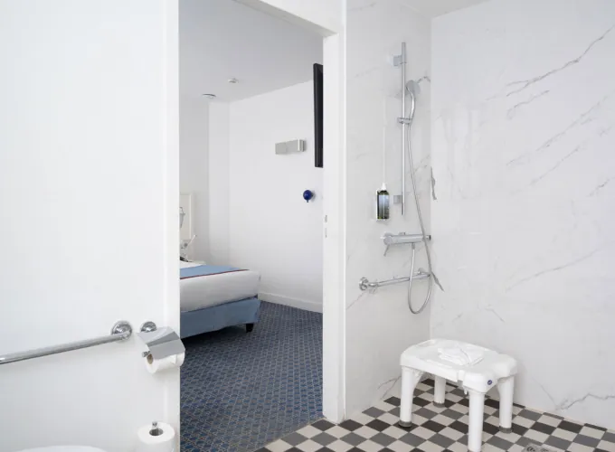 salle de bain accessible avec douche à l’italienne, siège, barres d’appui et vue sur la chambre à l’hôtel 34b