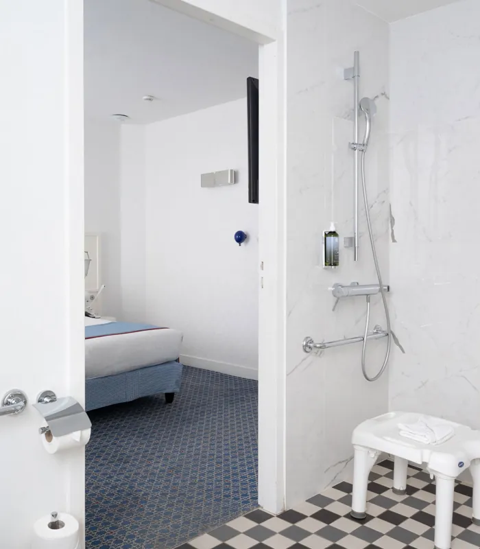 salle de bain accessible avec douche à l’italienne, siège, barres d’appui et vue sur la chambre à l’hôtel 34b