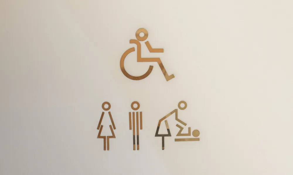 Pictogrammes dorés sur fond clair à l’Hôtel Astra Opéra représentant un fauteuil roulant, des toilettes mixtes et un espace de change bébé, sans braille.