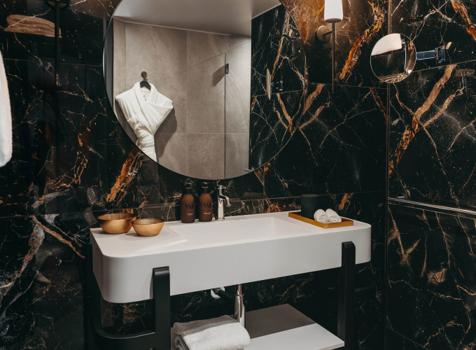 Salle de bains de la Chambre Deluxe de l’Hôtel Barnett à Paris avec marbre noir veiné, vasque blanche, miroir rond et douche.