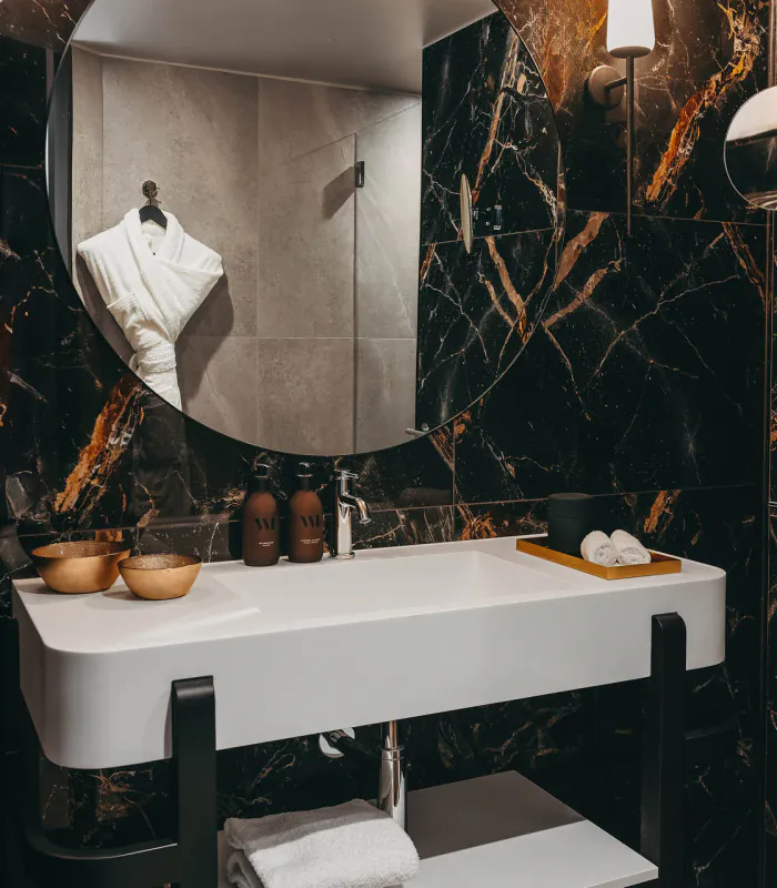 Salle de bains de la Chambre Deluxe de l’Hôtel Barnett à Paris avec marbre noir veiné, vasque blanche, miroir rond et douche.
