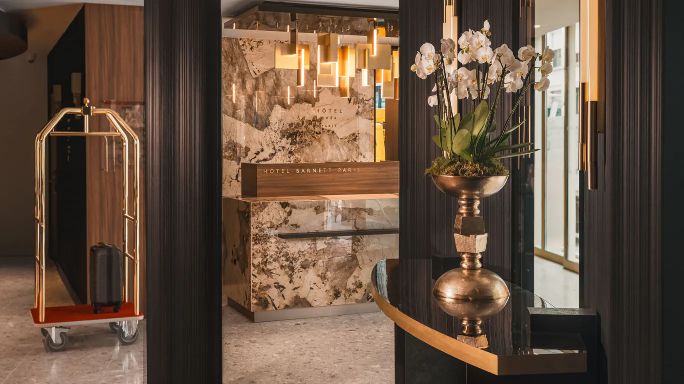 Réception de l’Hôtel Barnett à Paris avec comptoir en pierre, décoration florale et chariot à bagages.